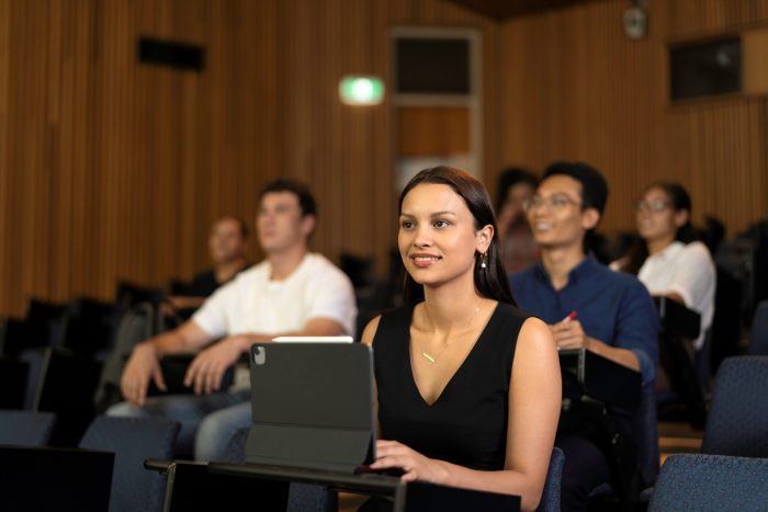 People seated in a lecture hall, with one person in the foreground using a tablet on a desk and others behind taking notes.
